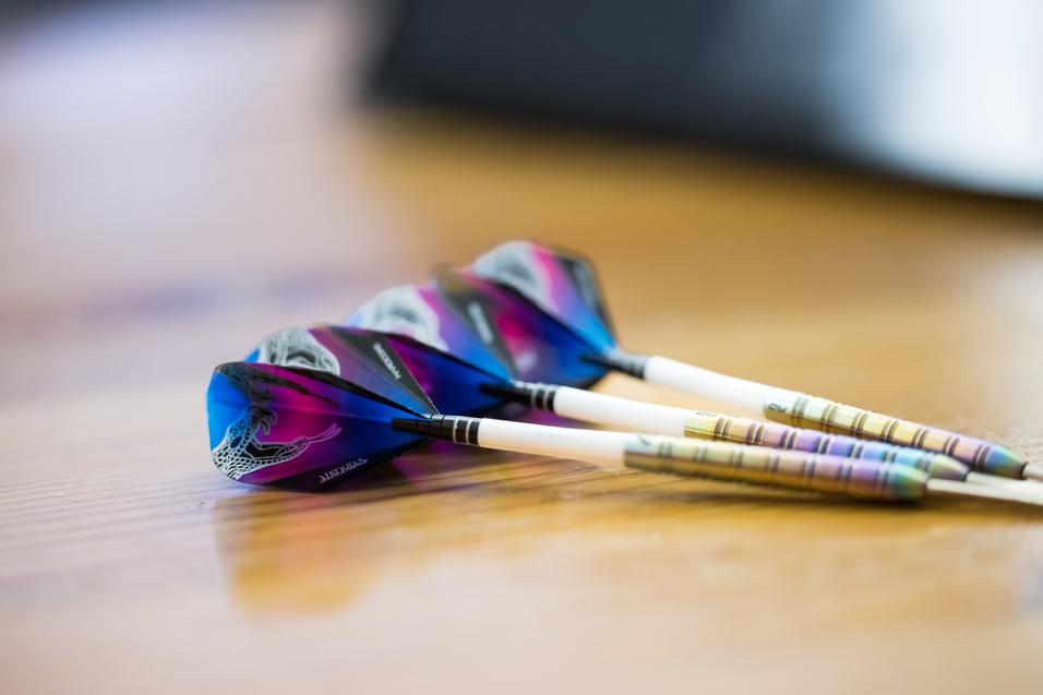 Drei Dartpfeile mit bunten Flights liegen auf einem Holztisch.