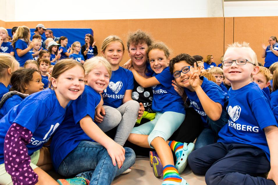 Gruppenkinder in blauen T-Shirts umarmen eine Frau, die lächelt, in einer Sporthalle voller Menschen.