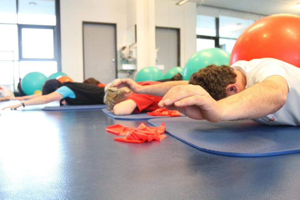 Gruppentraining mit Personen liegend auf Matten, während Bälle im Hintergrund sichtbar sind.