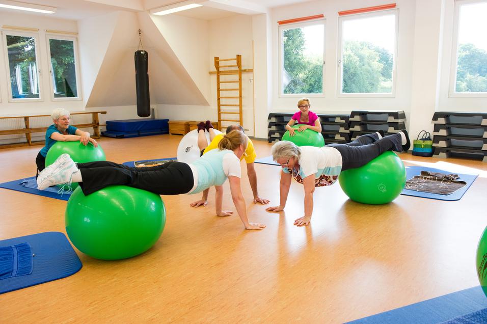 Gruppentraining auf Gymnastikbällen: Frauen führen Übungen in einem hellen Raum mit Holzboden und weißen Wänden aus.