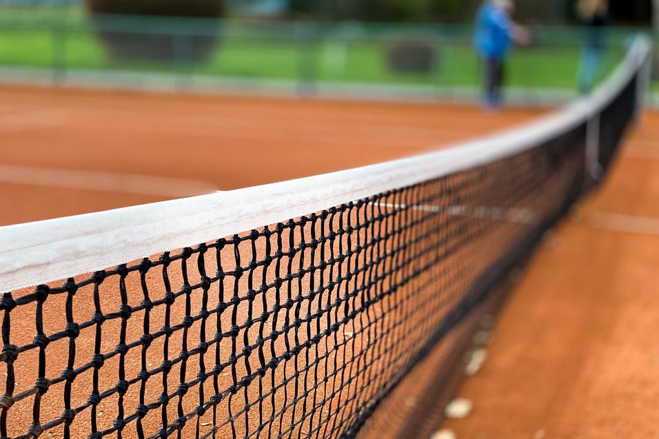 Nahaufnahme eines Tennisnetzes auf einem roten Sandplatz, Hintergrund unscharf mit grünen Bäumen.