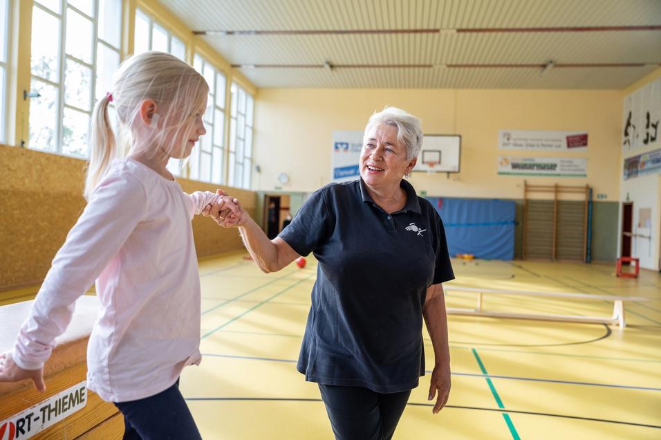 Ein junges Mädchen hält die Hand einer älteren Frau in einer Sporthalle mit bunten Sportgeräten im Hintergrund.